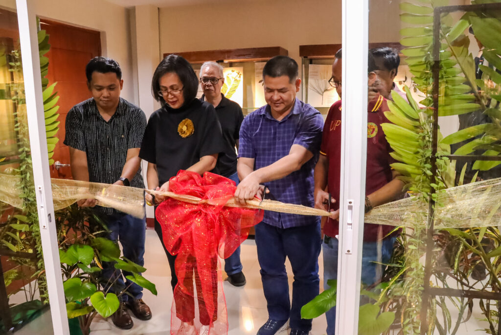 A group of officials gather at an entrance to perform a ceremonial ribbon-cutting, using scissors to slice through a wide, golden mesh ribbon decorated with a large red bow. Captured from just outside the doorway, the team works together to officially open the venue as onlookers watch.