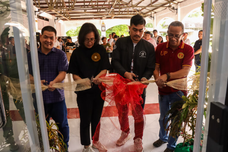 Four individuals participate in a ribbon-cutting ceremony at the entrance of an indoor venue, cutting a decorative ribbon with a large red bow. Guests are gathered in the background, witnessing the opening event.
