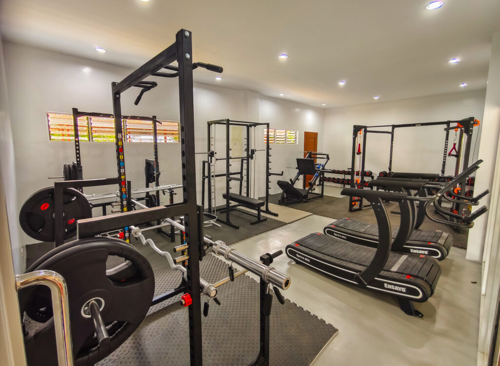 A well-lit indoor gym features various fitness equipment, including treadmills, squat racks, weight benches, barbells, and free weights arranged neatly across the room. The clean exercise space is fitted with rubber flooring and bright overhead lighting.