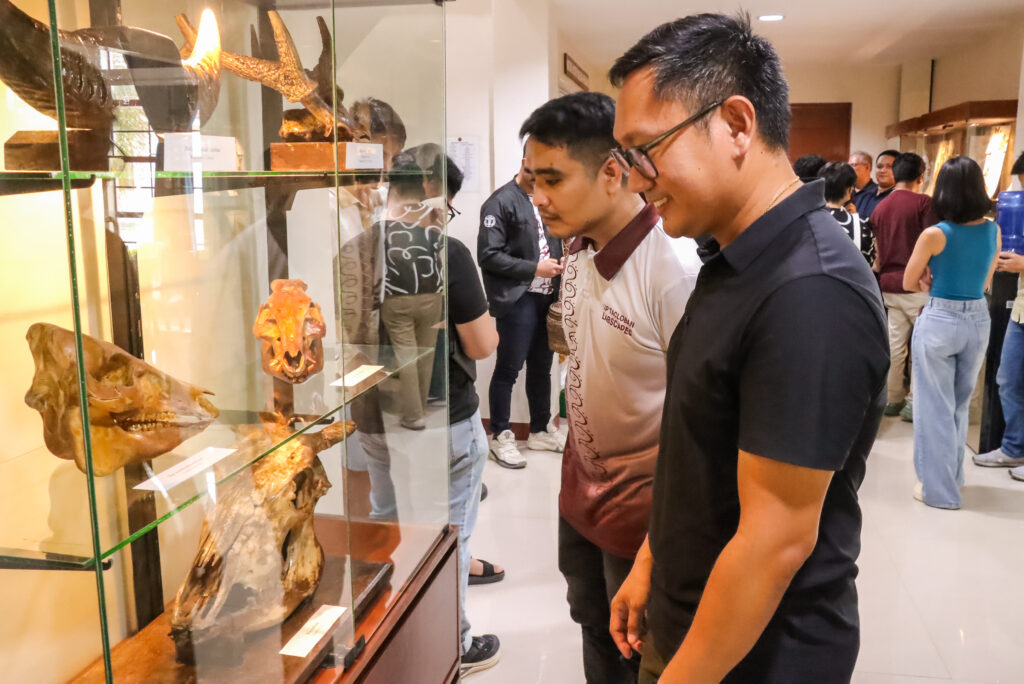 Two men stand side by side, smiling as they look into a glass museum display case containing various animal skulls and horns. Other visitors are visible in the background browsing the gallery.