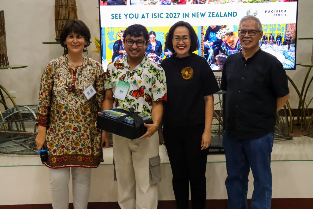 ALT Text: Four smiling individuals stand shoulder-to-shoulder in front of a presentation screen that reads "SEE YOU AT ISIC 2027 IN NEW ZEALAND." The second man from the left, wearing a colorful butterfly-patterned shirt, holds a black gift box containing a book, flanked by three other colleagues posing for the photo.