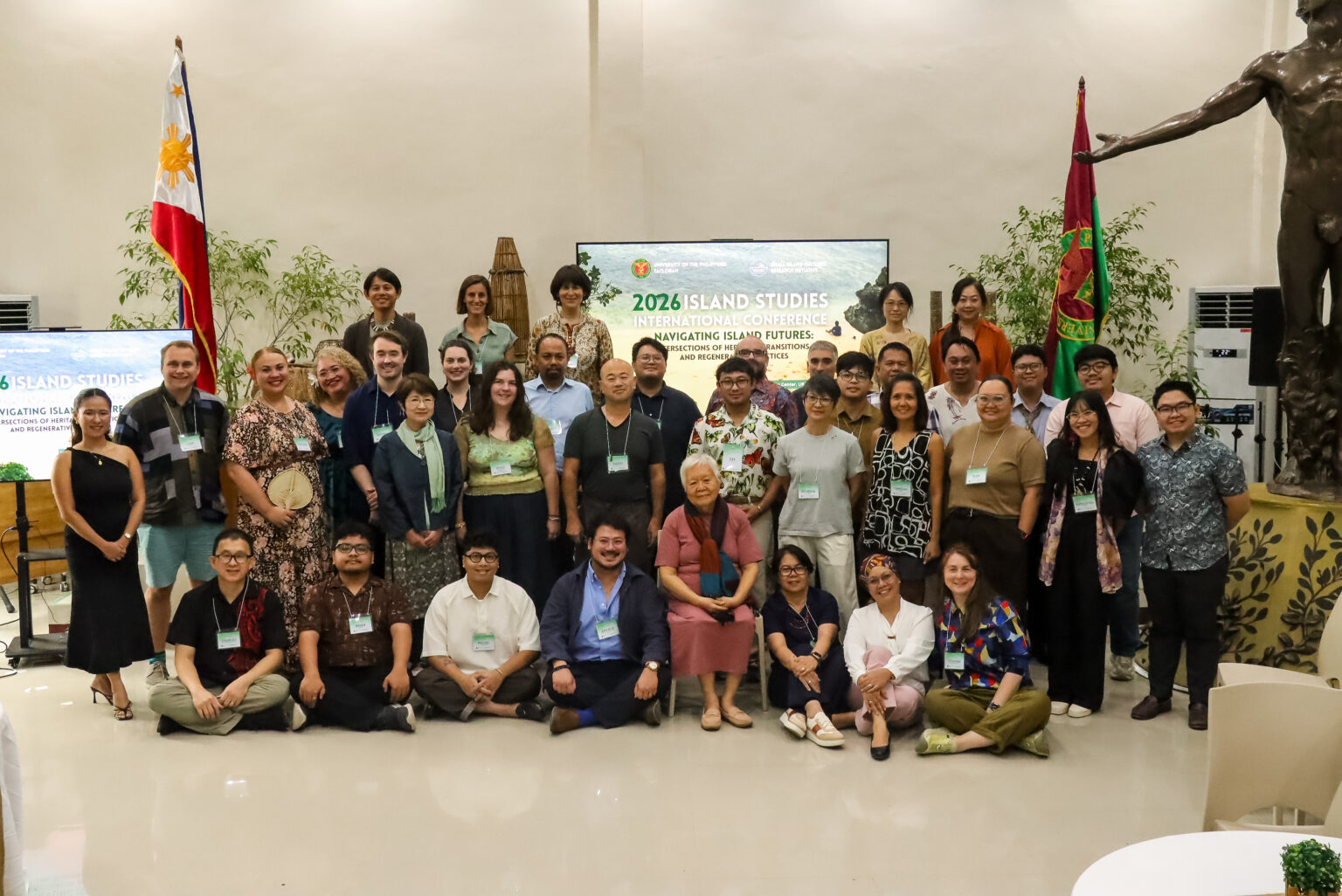A group photo of approximately 50 participants of the 2026 Island Studies International Conference (ISIC) gathered inside the Leyte Samar Heritage Center in Tacloban City. The Philippine and UP flags are visible on either side of the group, with the iconic statue inside the venue partially seen on the right. The participants, wearing conference name tags, smile toward the camera at the conclusion of the three-day international conference.