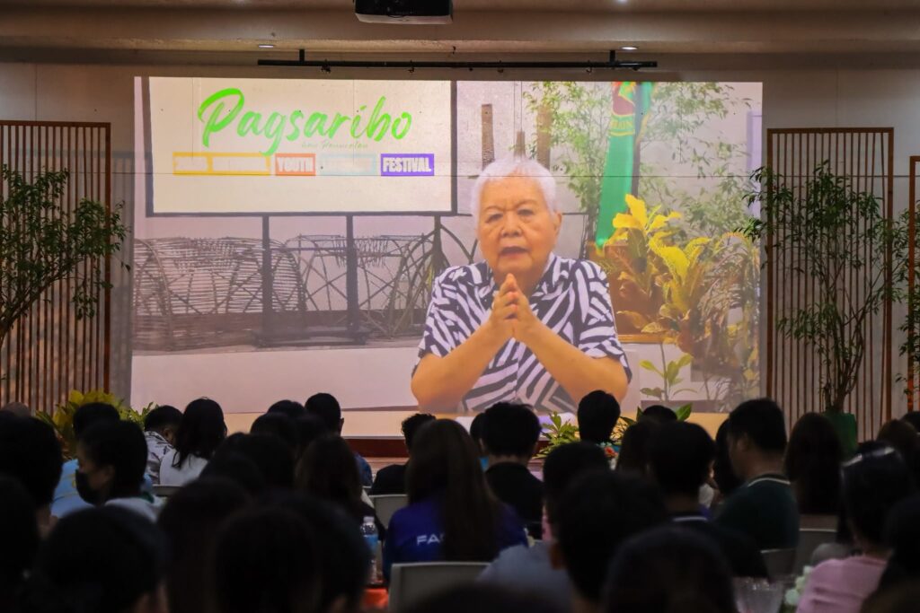 An audience watches a large projection screen displaying Professor Emeritus Merlie Alunan delivering her address virtually for the Pagsaribo han Panuratan National Youth Literary Festival.
