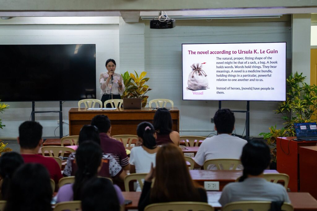 Writer Kaisa Aquino speaks into a microphone while standing in front of workshop slides about Ursula K. Le Guin's concept of the novel as a vessel, with participants listening in a classroom setting.