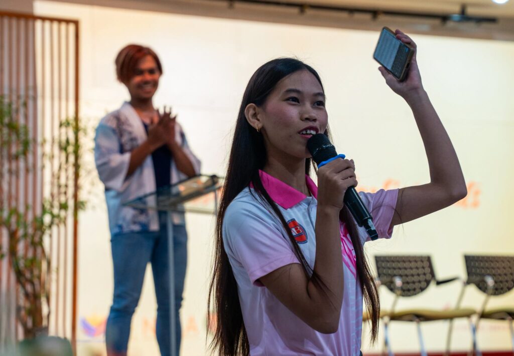 A young female participant in a pink polo shirt speaks into a microphone while holding up her smartphone on stage, as another person smiles and applauds in the background.
