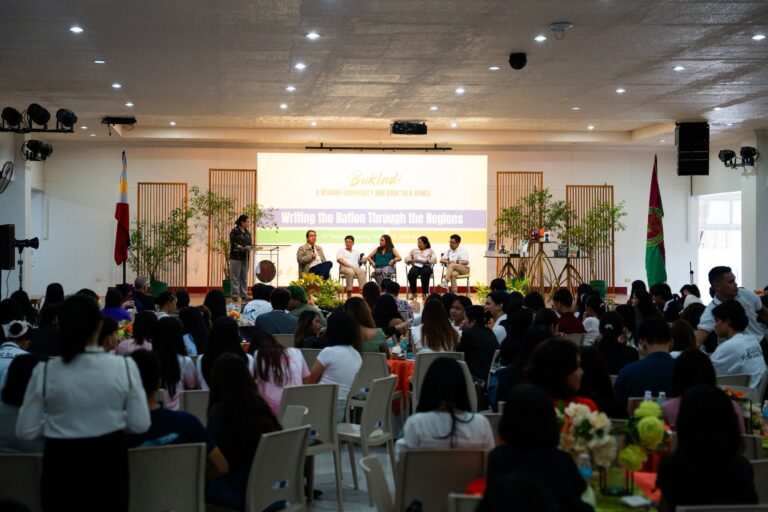 A wide shot of a crowded auditorium at UP Tacloban during a panel discussion on stage titled "Buklad: A Reading Community and Book Talk Series" under the theme "Writing the Nation Through the Regions."