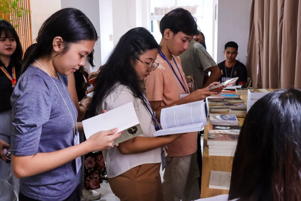 Several young festival attendees wearing identification lanyards stand in a line to browse and read through various books displayed on a long wooden table.