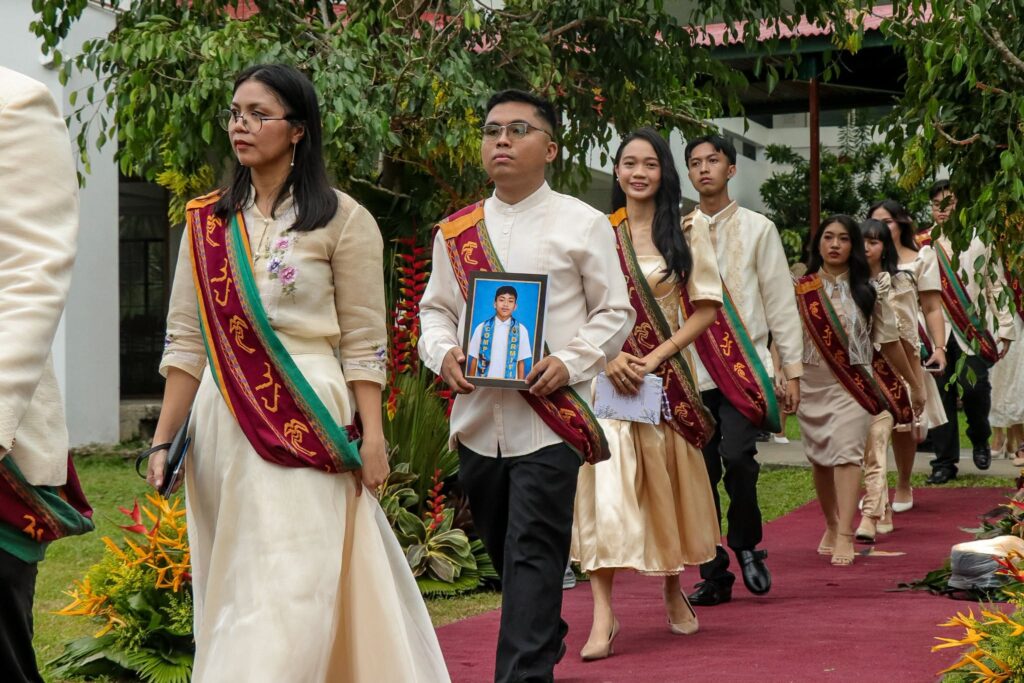 A line of UP Tacloban graduates dressed in formal attire and traditional Sablay sashes walk down a red carpet during an outdoor ceremony. In the center, a male graduate solemnly carries a framed portrait of his younger brother, honoring their memory during the processional.