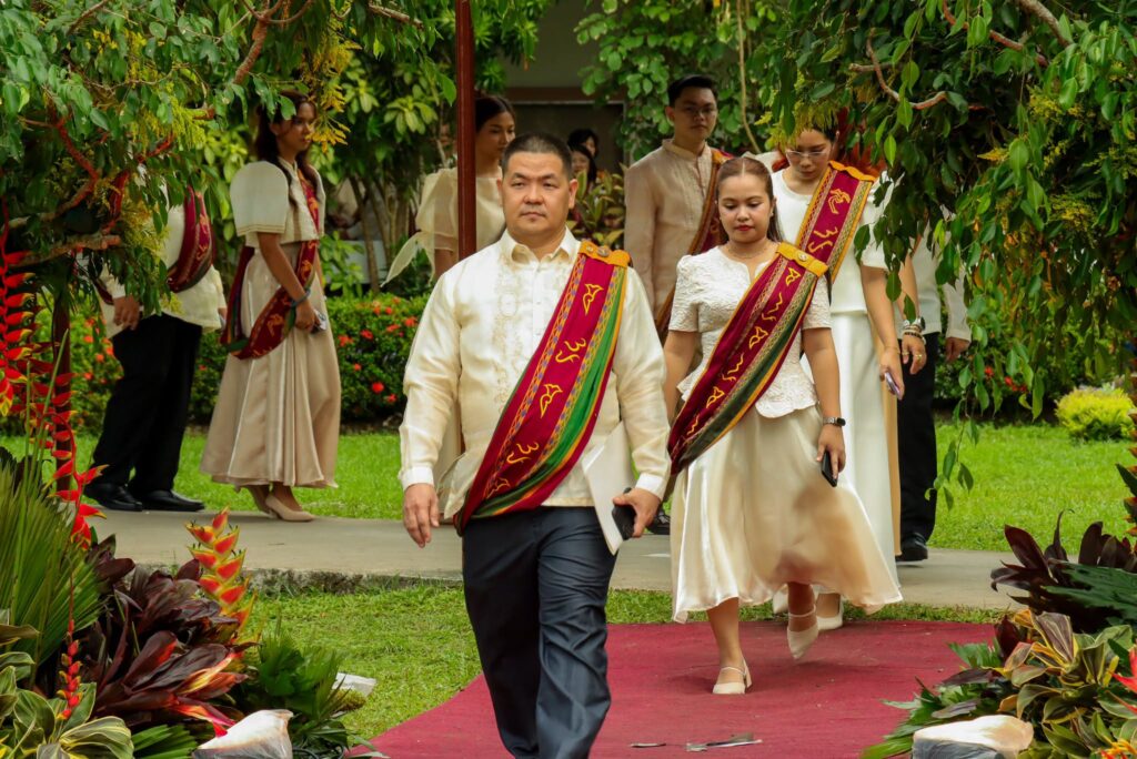 A group of university faculty and officials walks down a red carpet in an outdoor setting during a formal academic procession. They are dressed in formal attire, including cream-colored barongs and dresses, accented with the maroon and green University of the Philippines Sablay sash.