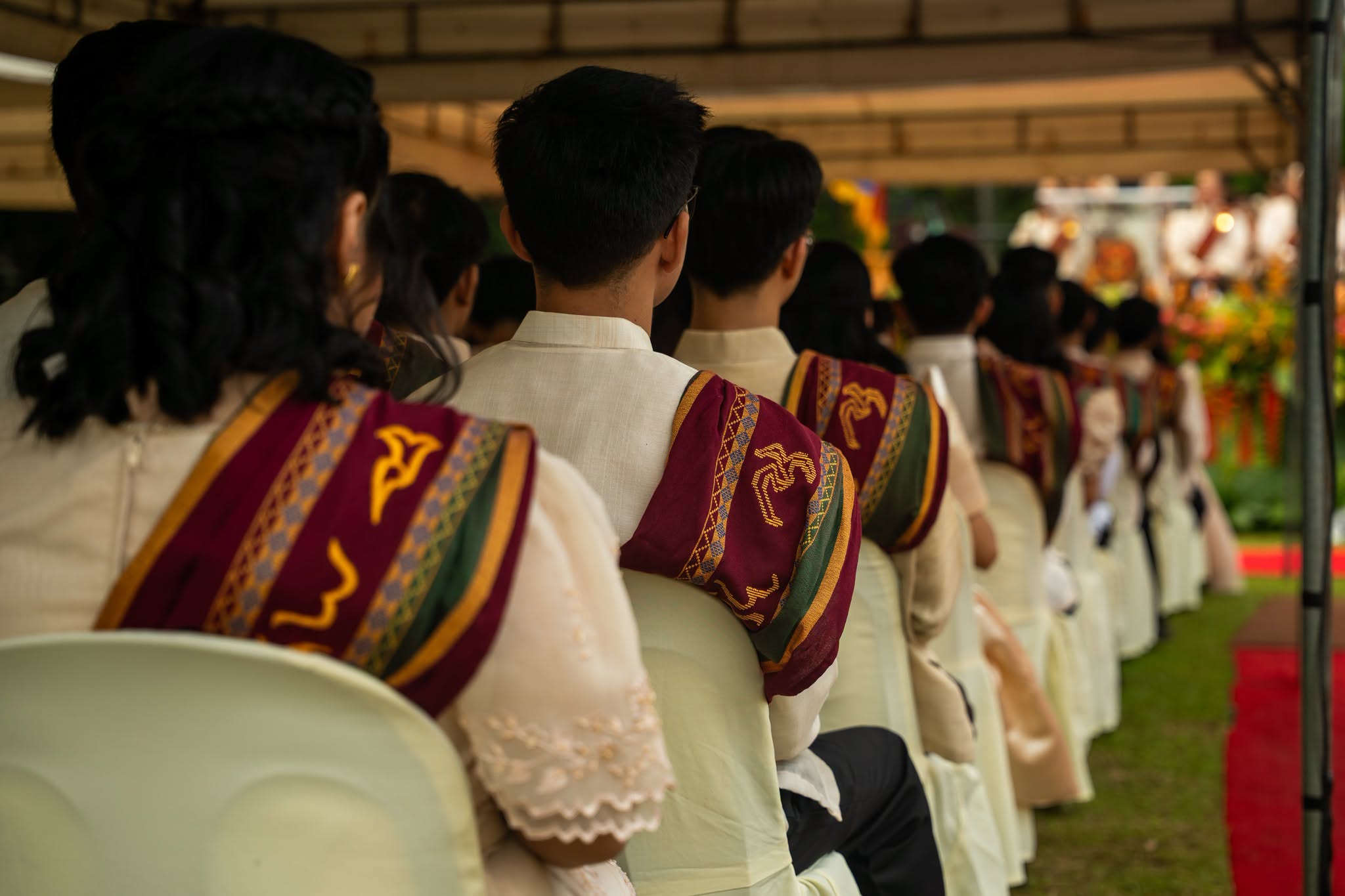A row of graduates is seated under an outdoor tent, captured from behind during a commencement ceremony. They are dressed in formal cream-colored attire with the traditional University of the Philippines maroon and green "Sablay" sash draped over their shoulders.