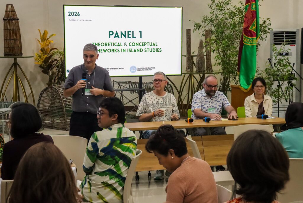 A speaker stands holding a microphone in front of a presentation screen that reads "Panel 1: Theoretical & Conceptual Frameworks in Island Studies" at a 2026 conference in Tacloban City, Philippines. Three other panelists sit at a wooden table beside him, while attendees listen in the foreground.