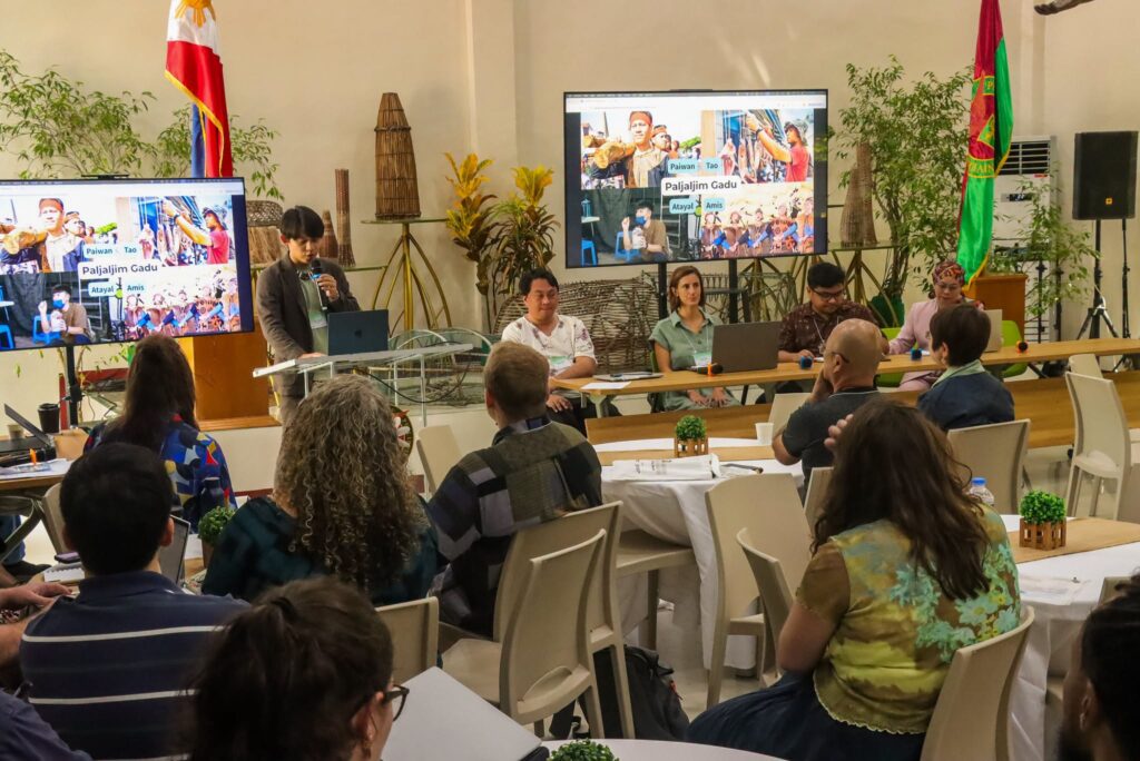 A presenter stands at a podium speaking into a microphone during an academic session, with a laptop open in front of him. Behind him, a large display screen shows a presentation slide titled "Paljaljim Gadu" featuring images of indigenous groups (labeled Paiwan, Tao, Atayal, and Amis). Four panelists sit at a long table adjacent to the screen, while attendees sit at round tables in the foreground listening to the presentation.