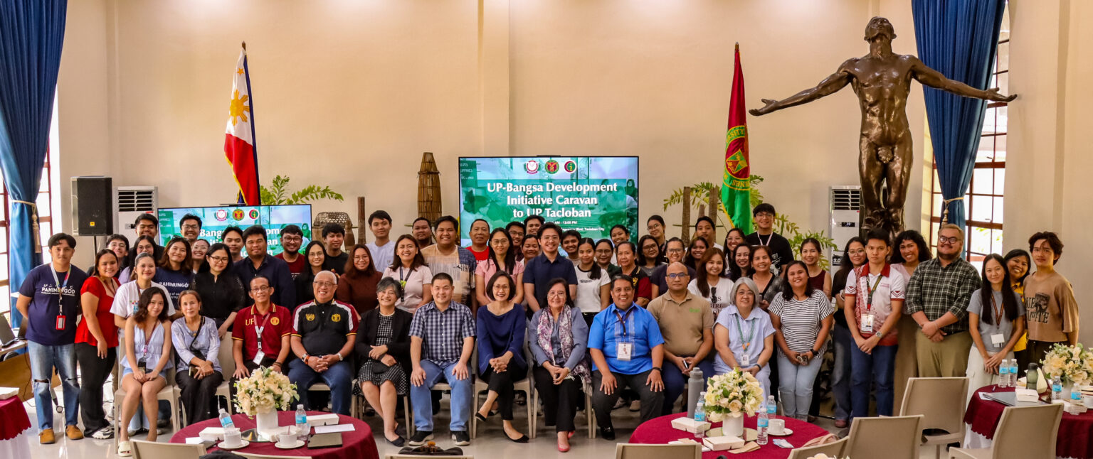 A large group pose featuring event attendees, speakers, and organizers inside a high-ceiling hall. In the background, a digital screen displays "UP-Bangsa Development Initiative Caravan to UP Tacloban," flanked by the Philippine flag, the UP flag, and the iconic Oblation statue.