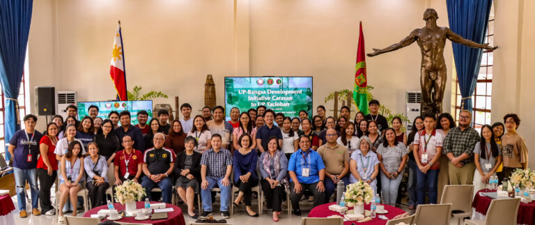 A large group pose featuring event attendees, speakers, and organizers inside a high-ceiling hall. In the background, a digital screen displays "UP-Bangsa Development Initiative Caravan to UP Tacloban," flanked by the Philippine flag, the UP flag, and the iconic Oblation statue.