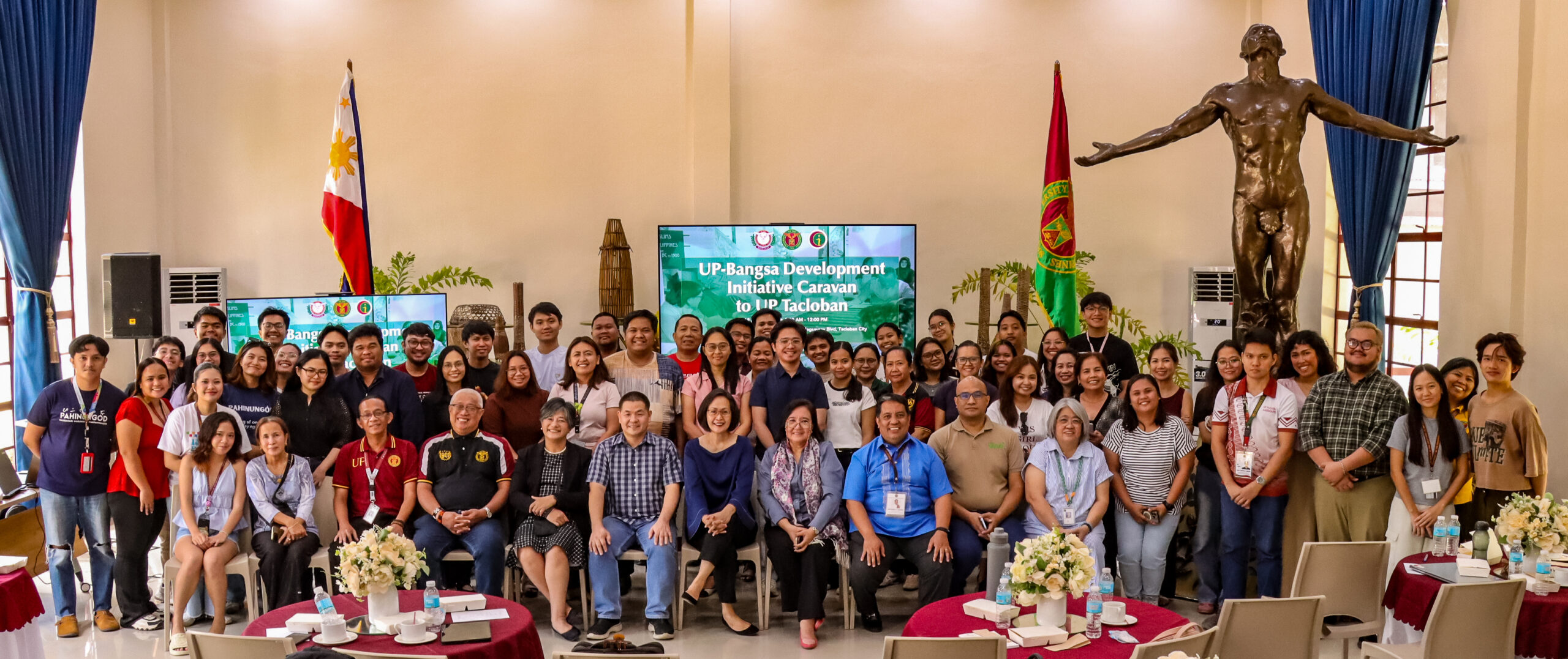 A large group pose featuring event attendees, speakers, and organizers inside a high-ceiling hall. In the background, a digital screen displays "UP-Bangsa Development Initiative Caravan to UP Tacloban," flanked by the Philippine flag, the UP flag, and the iconic Oblation statue.