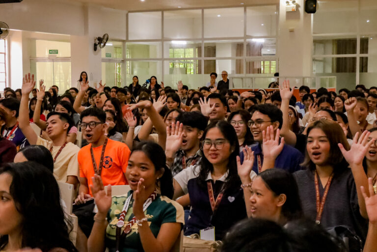 A large group of UP Tacloban students seated inside an orientation venue, with many raising their hands and smiling as they participate in an interactive activity.