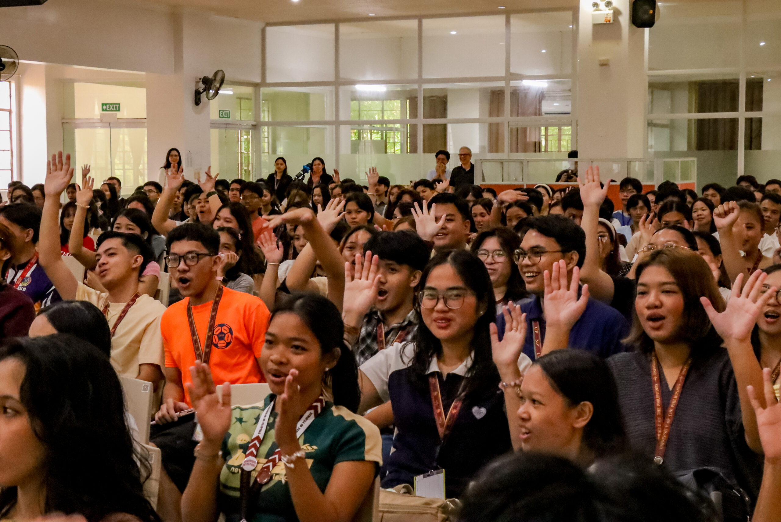 A large group of UP Tacloban students seated inside an orientation venue, with many raising their hands and smiling as they participate in an interactive activity.