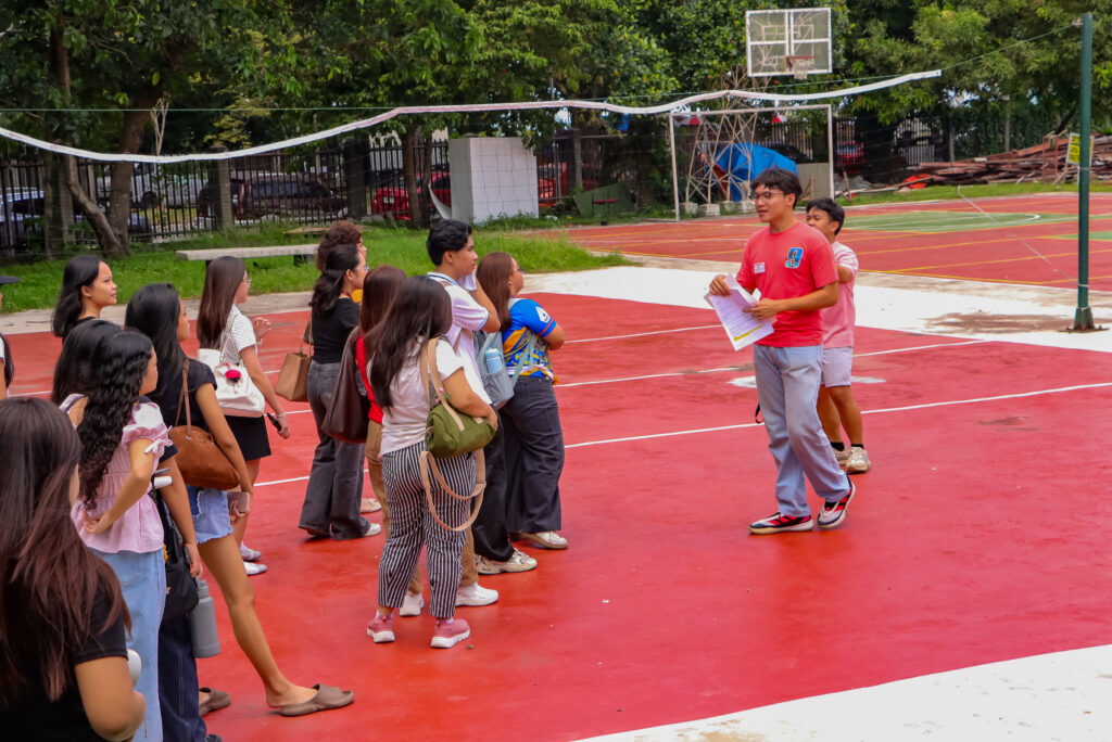 A male tour guide in a pink t-shirt speaks to a group of students gathered on a red outdoor basketball court.