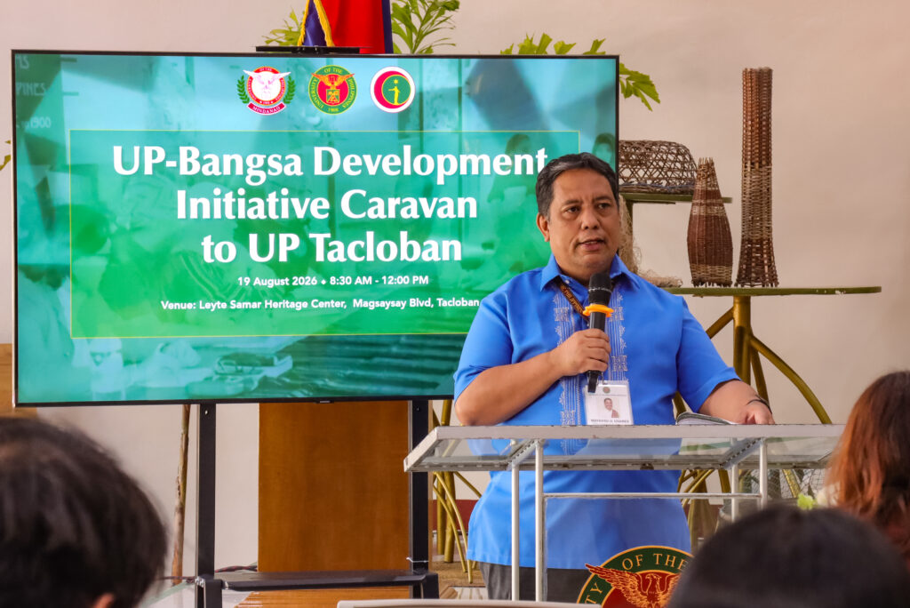 A man in a blue Barong Tagalog speaking at a podium with a microphone. Next to him, a digital display screen reads "UP-Bangsa Development Initiative Caravan to UP Tacloban."