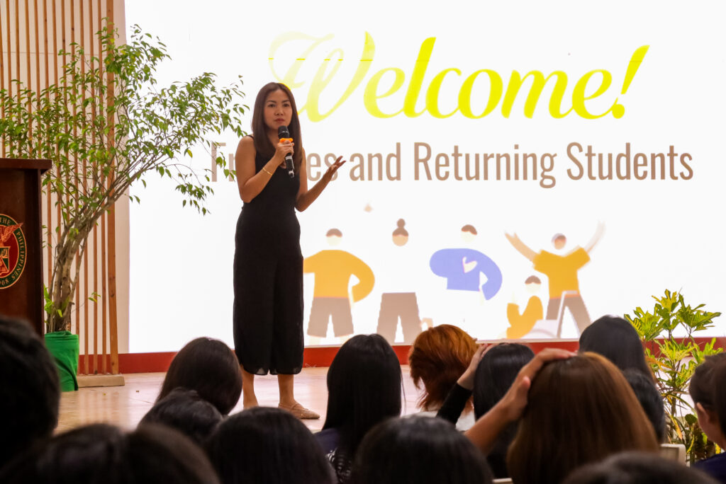 A woman in a black dress speaks into a microphone on stage during the CSSM orientation, with students seated in the audience and a welcome presentation displayed on the screen behind her.