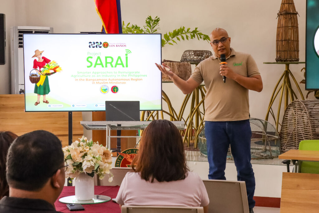 A speaker in a beige polo shirt gestures while presenting next to a digital display for "Project SARAI: Smarter Approaches to Reinvigorate Agriculture as an Industry in the Philippines."