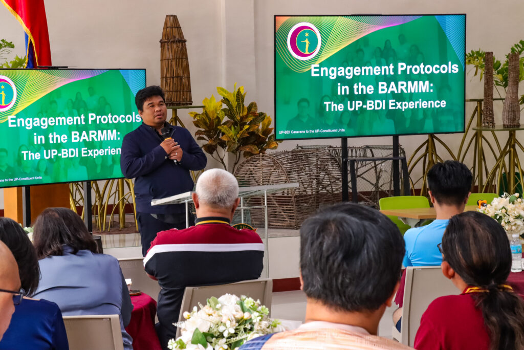 A speaker addresses the audience from a podium, standing in front of two screens displaying a slide titled "Engagement Protocols in the BARMM: The UP-BDI Experience."