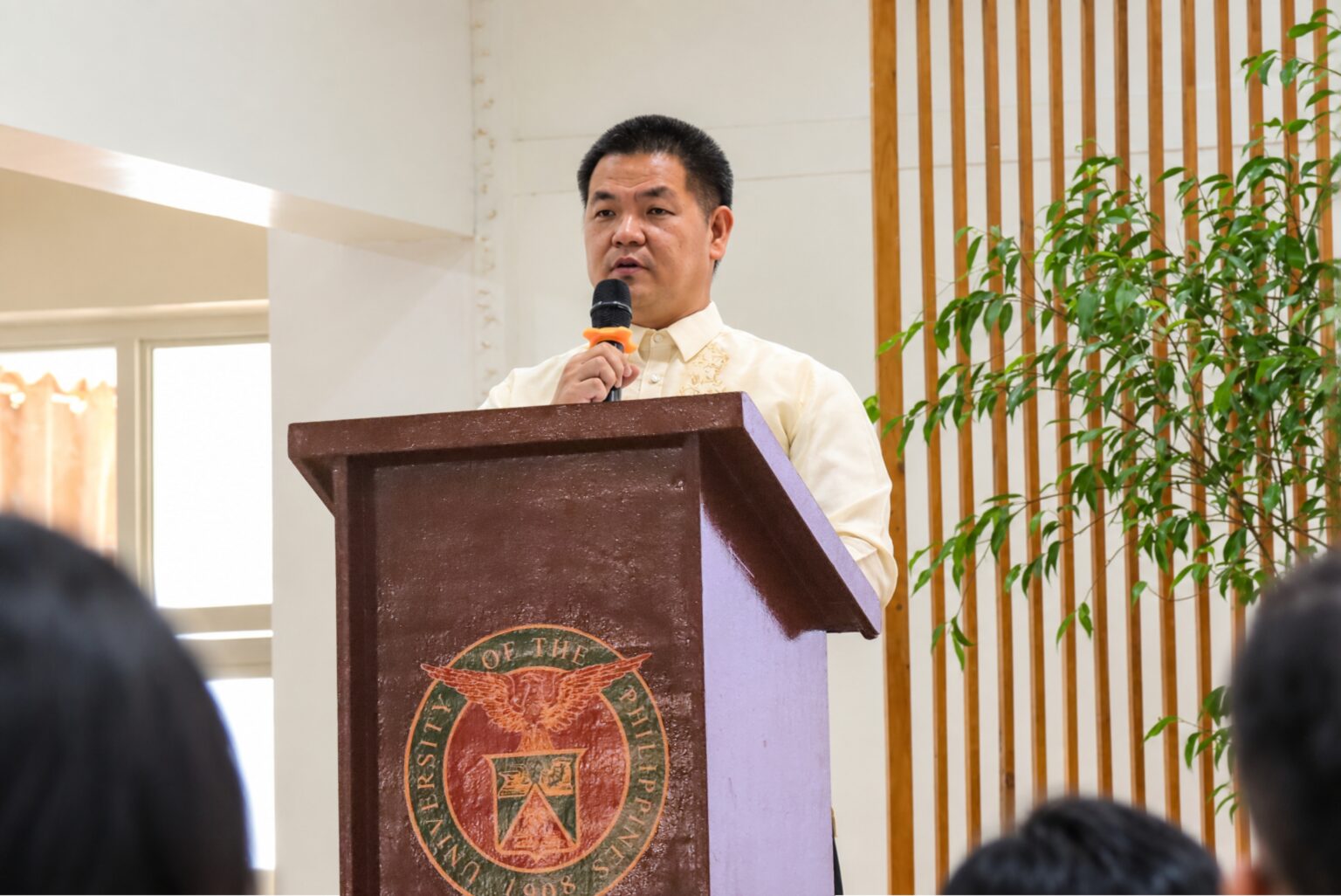 A man in a cream Barong Tagalog stands at a dark wooden wooden podium featuring the University of the Philippines seal, speaking into a microphone while addressing an audience.