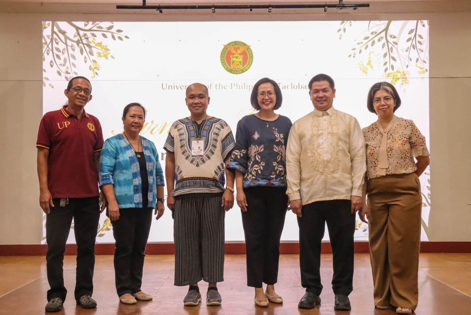 Six university officials stand in a row on stage smiling for a group photo in front of a backdrop displaying the University of the Philippines seal.