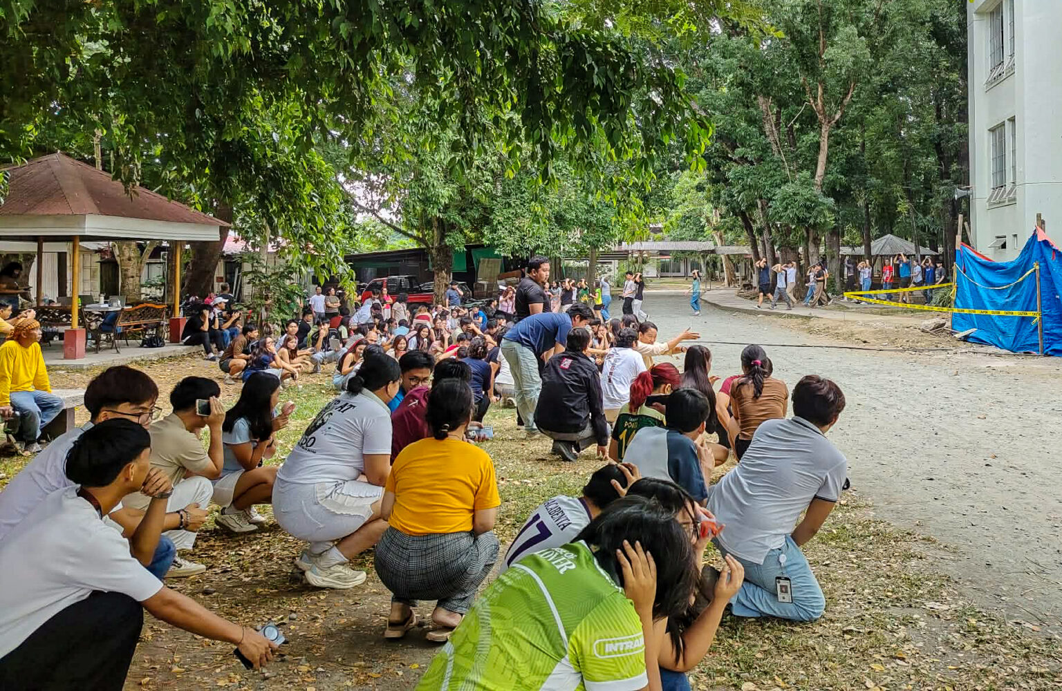 A large group of students gather outdoors on a tree-lined campus ground, crouching and kneeling while covering their heads during an emergency preparedness drill.