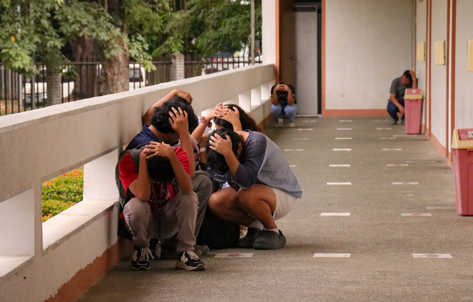 Alt Text A group of students crouch along an outdoor covered hallway, covering their heads with both hands against a concrete ledge during an earthquake drill.