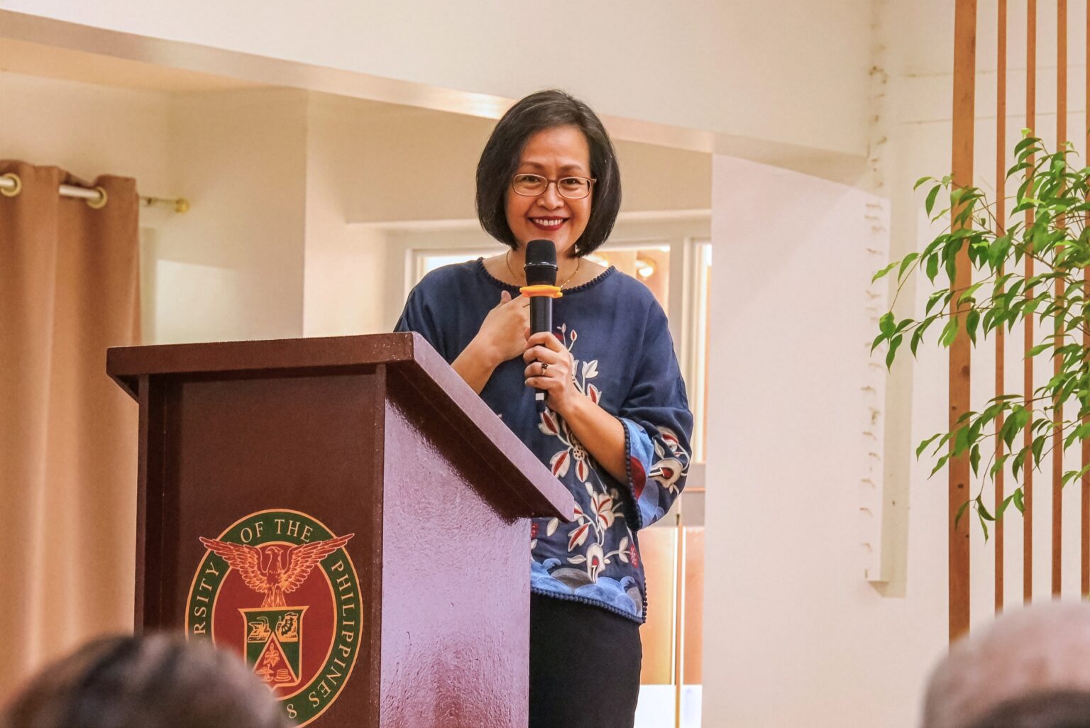 A woman wearing glasses and an embroidered blue top stands behind a dark wooden podium bearing the University of the Philippines seal, holding a microphone in one hand with her other hand resting over her heart as she smiles and addresses the audience.