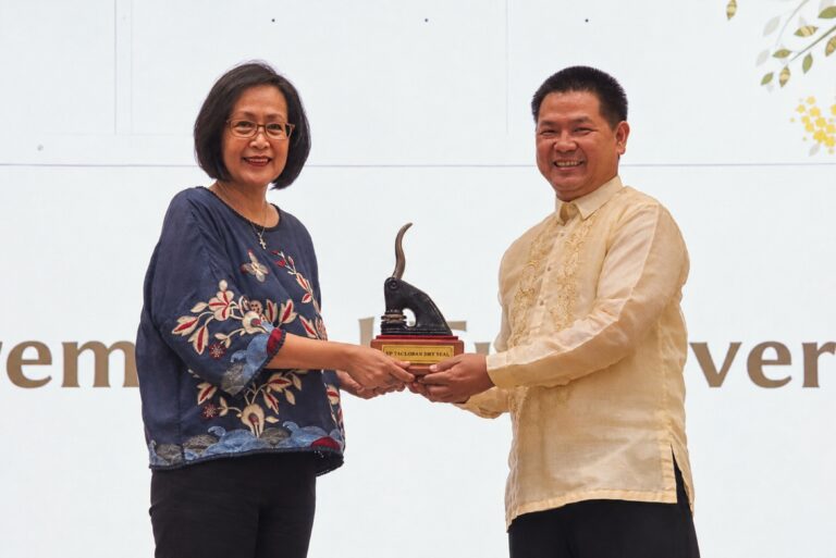 A woman in a embroidered blue blouse and a man in a beige Barong Tagalog smile at the camera while jointly holding an award trophy shaped like a dry seal embosser on a wooden base.